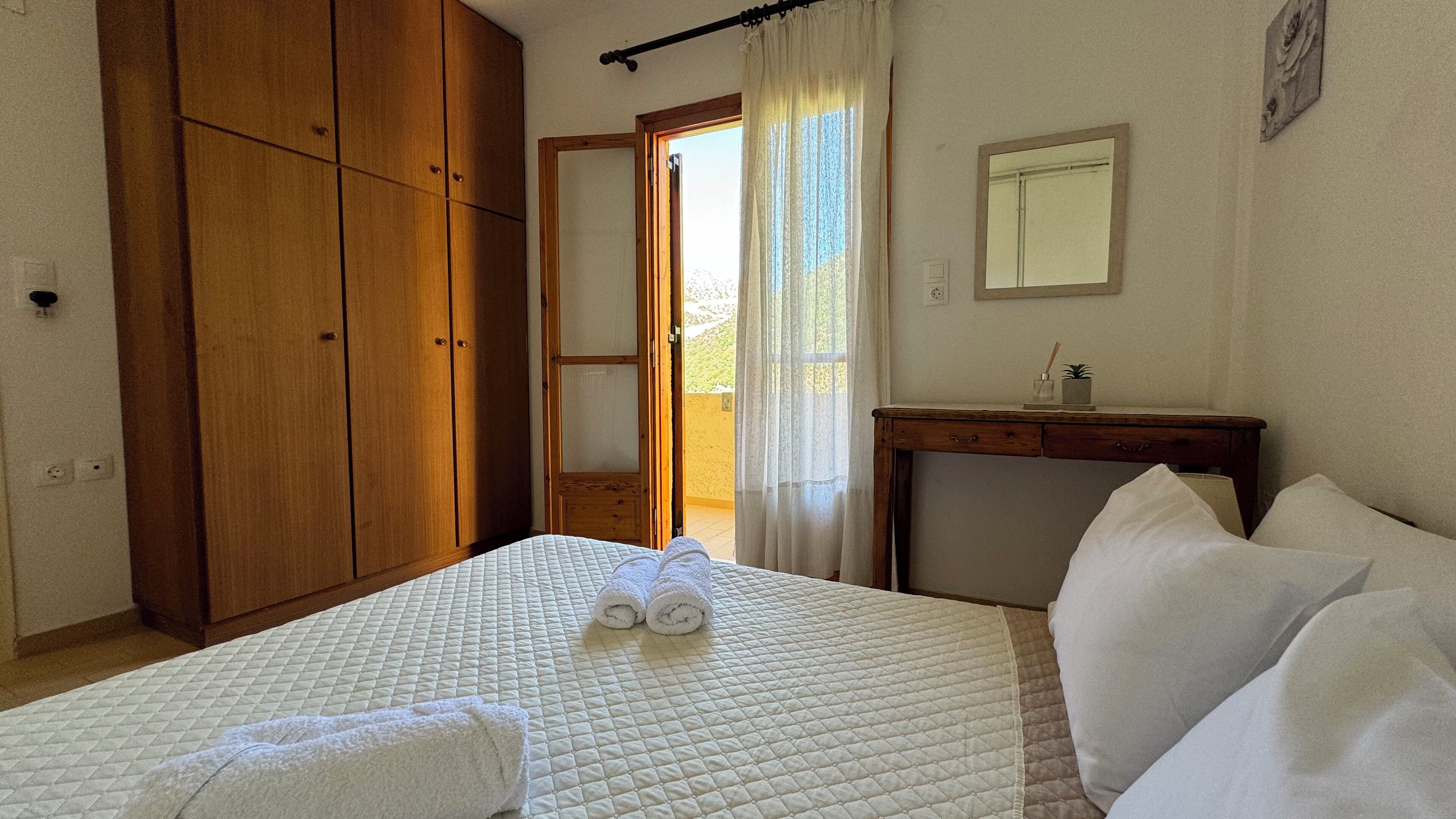 A bedroom with a bed, a wooden wardrobe, a desk with a mirror, and a window with curtains.