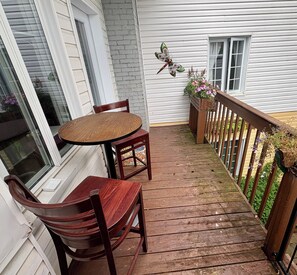 Une terrasse en bois avec une table ronde et deux chaises, un mur en briques, une libellule décorative et une balustrade en bois ornée de fleurs.