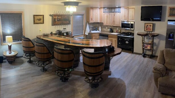A modern kitchen with a central island, bar stools, wooden cabinets, a microwave, and a flat-screen TV. - Whiskey Barrel Bunkhouse (Sunrise Beach)
