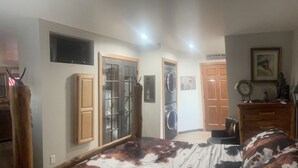 A bedroom with a bed, a wooden wardrobe, a washing machine, and a television on the wall. - Whiskey Barrel Bunkhouse (Sunrise Beach)