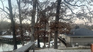 A gravel patio with a wooden bench, trees, and a view of a water body and buildings in the background. - Whiskey Barrel Bunkhouse (Sunrise Beach)
