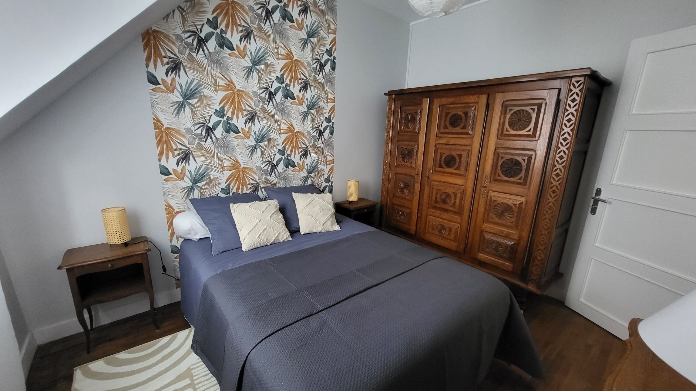 A bedroom with a bed, a wardrobe, a nightstand, and a wall with a tropical leaf pattern.