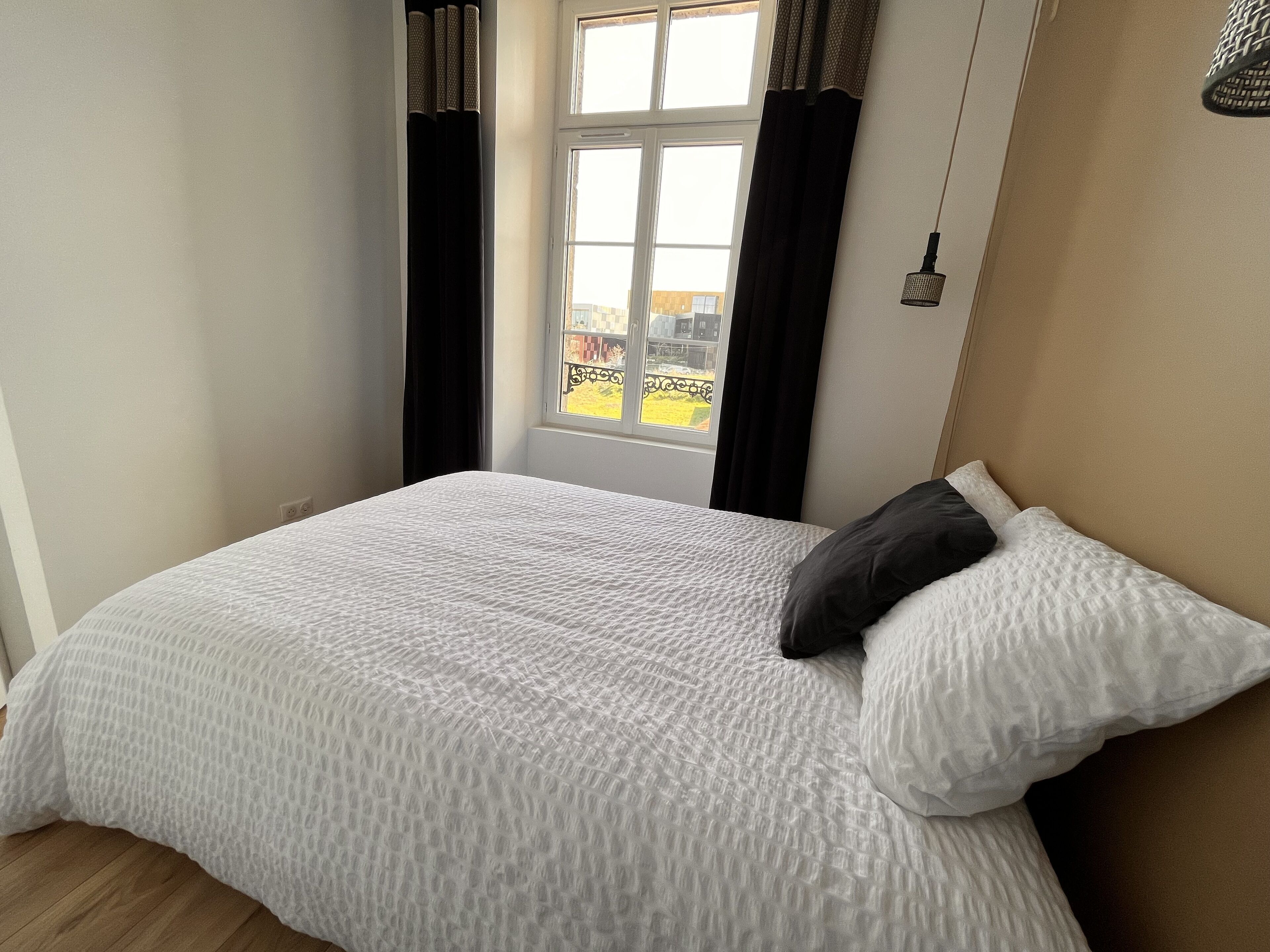 Une chambre avec un grand lit, une fenêtre avec des rideaux et une vue sur un bâtiment à l’extérieur.
