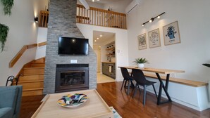 A modern living room with a fireplace, a TV mounted on a brick wall, a dining area with a table and chairs, and a staircase leading to an upper level. - Central & Cozy Lakeview condo within walking distance of Lake Memphremagog. (Magog)