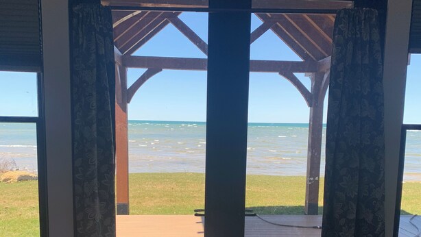 Una habitación con cama, una terraza de madera y vista a la playa y al océano a través de un ventanal con cortinas.