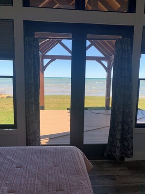 Una habitación con cama, una terraza de madera y vista a la playa y al océano a través de un ventanal con cortinas.
