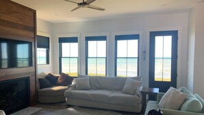 Una sala de estar con chimenea, ventilador de techo y grandes ventanales con vista al mar.