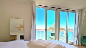 Une chambre d’hôtel avec un lit, un ventilateur de plafond, une grande fenêtre donnant sur la mer et une chaise près de la fenêtre.