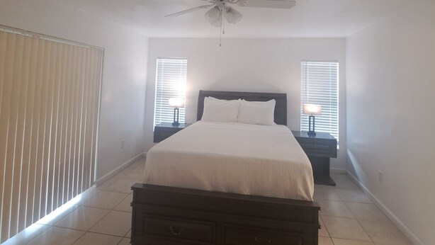 A bedroom with a bed, a ceiling fan, two bedside lamps, and a window with blinds.