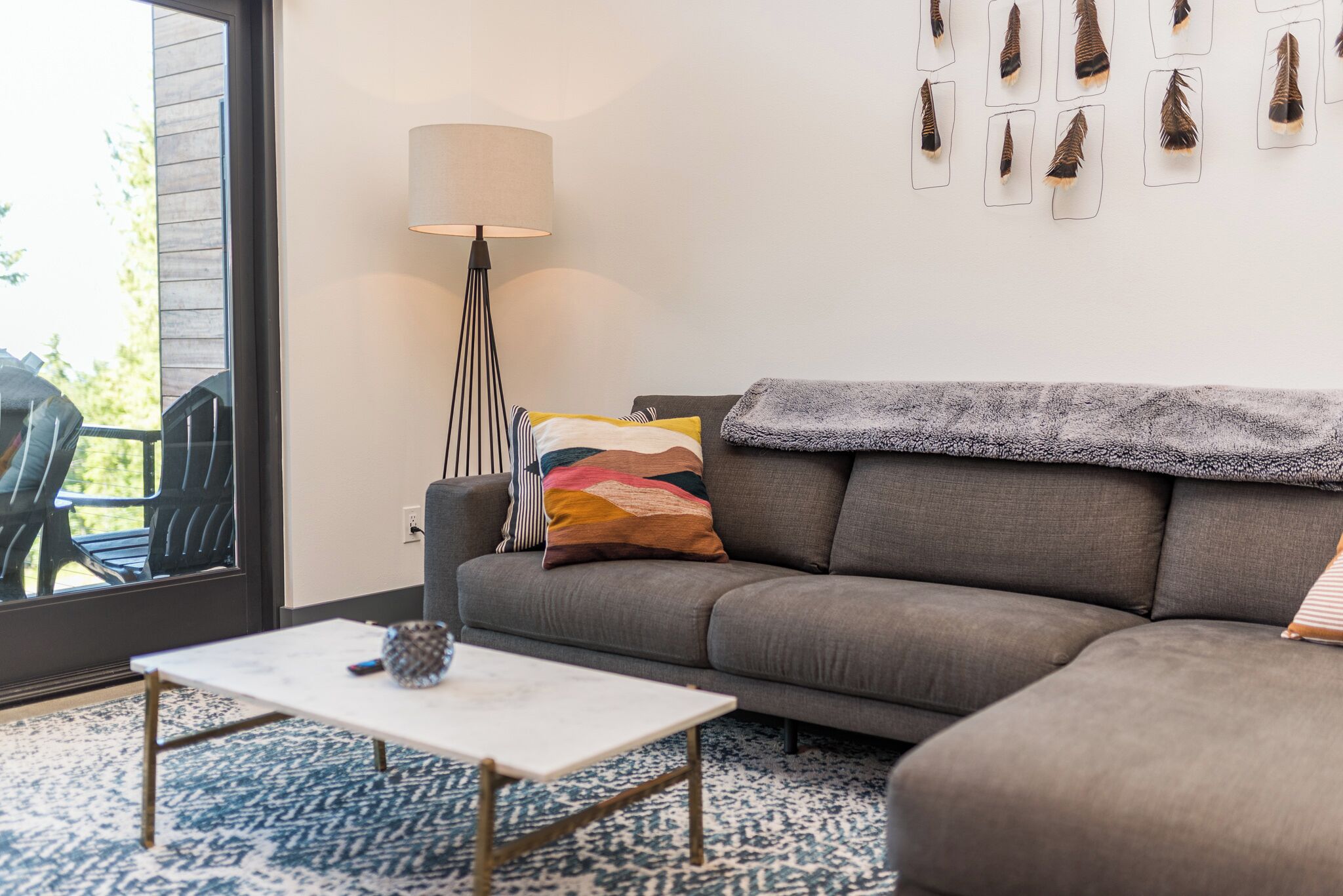 A modern living room with a grey sofa, a white coffee table, a floor lamp, and a large window with a view of outdoor chairs.