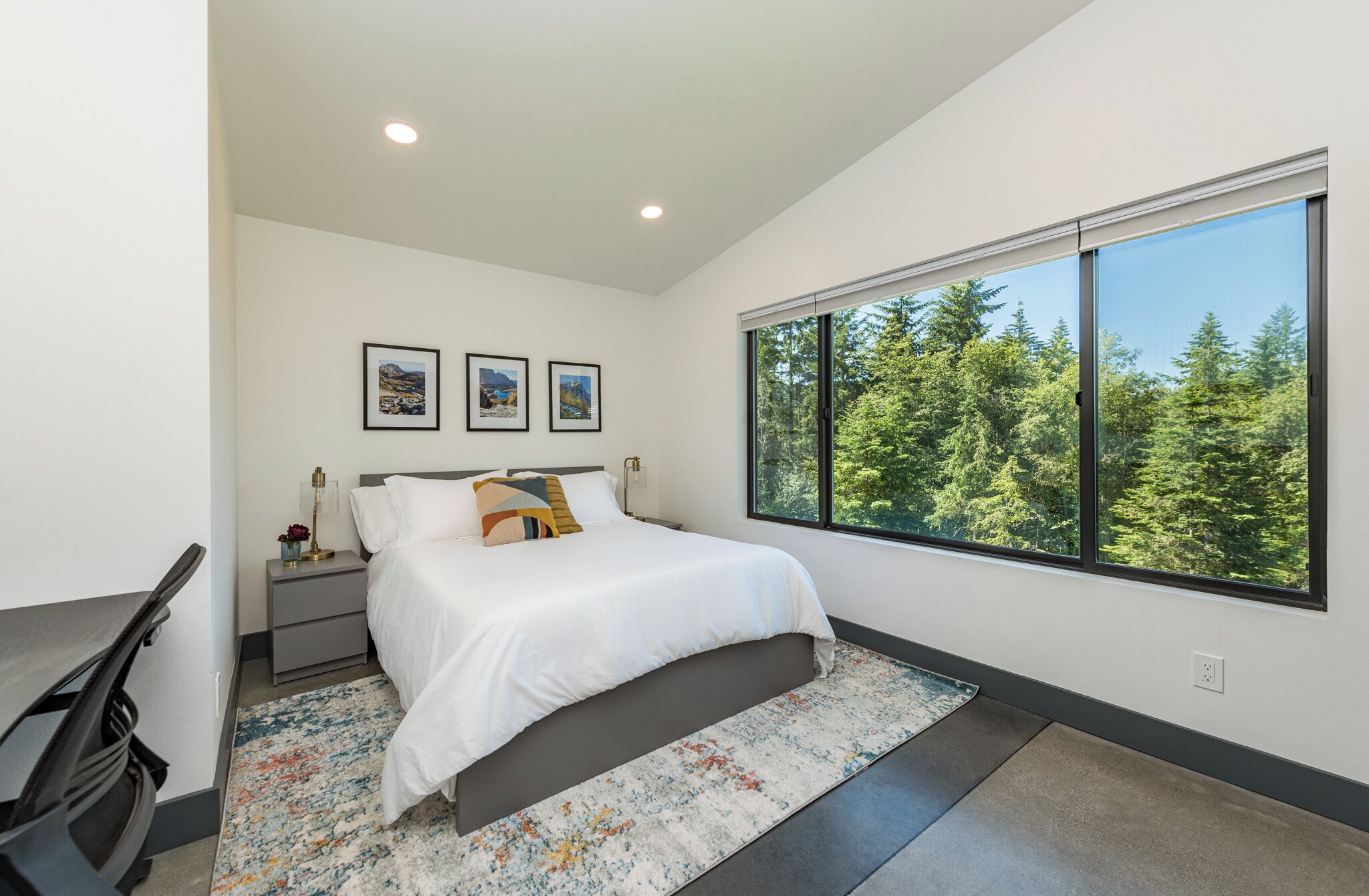A modern bedroom with a large bed, a desk, and a window with a forest view.