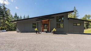 A modern two-story house with a sloped roof, large windows, and a wooden front door. - Custom modern home near Olympic National Park (Port Angeles)