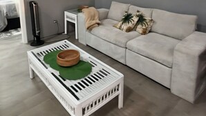 Un salón con un sofá blanco, una mesa de centro blanca con un posavasos verde, un cuadro en la pared y un radiador.