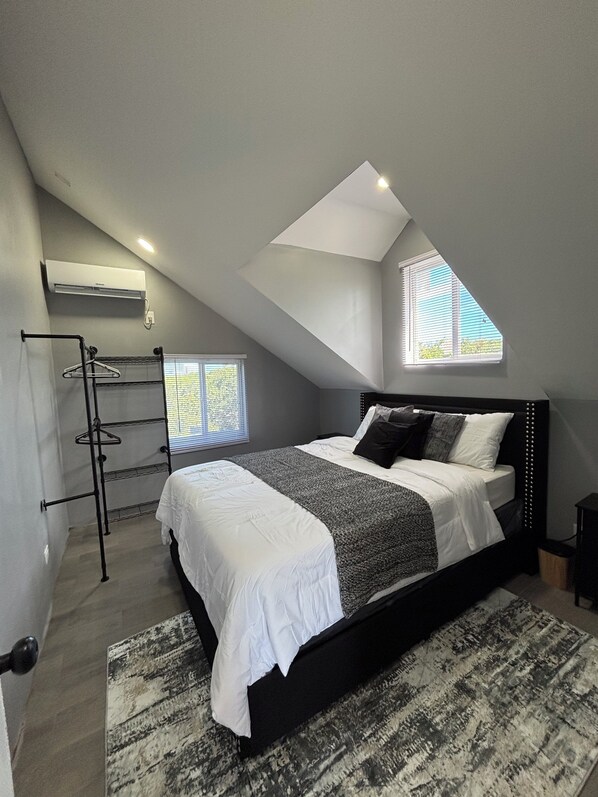 A bedroom with a sloped ceiling, a bed with a headboard, a hanging clothes rack, and a window with blinds.
