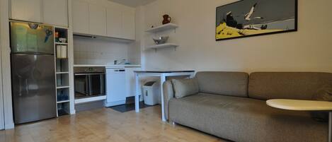 A modern kitchen with white cabinets, a built-in oven, and a stainless steel refrigerator. There is a grey sofa and a small table with a white tray in the living area.