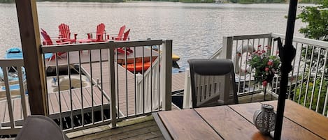 Een houten terras met buitenmeubels, met uitzicht op een meer en een huis op de achtergrond.
