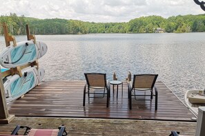 Une terrasse en bois avec deux chaises et une petite table, d’où l’on peut admirer un lac avec des bateaux et une maison au loin.