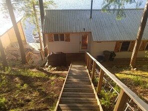 Une terrasse en bois mène à une petite cabane surmontée d’un toit en métal, qui est entourée d’arbres et bordée par un plan d’eau.