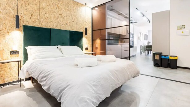 A modern hotel room with a large bed, wooden wall paneling, and a glass partition separating the bedroom from the kitchen area.
