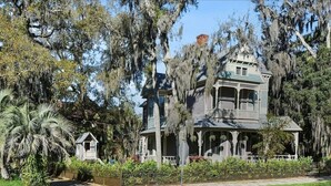 A large, historic house with a wrap-around porch, surrounded by lush greenery and trees.