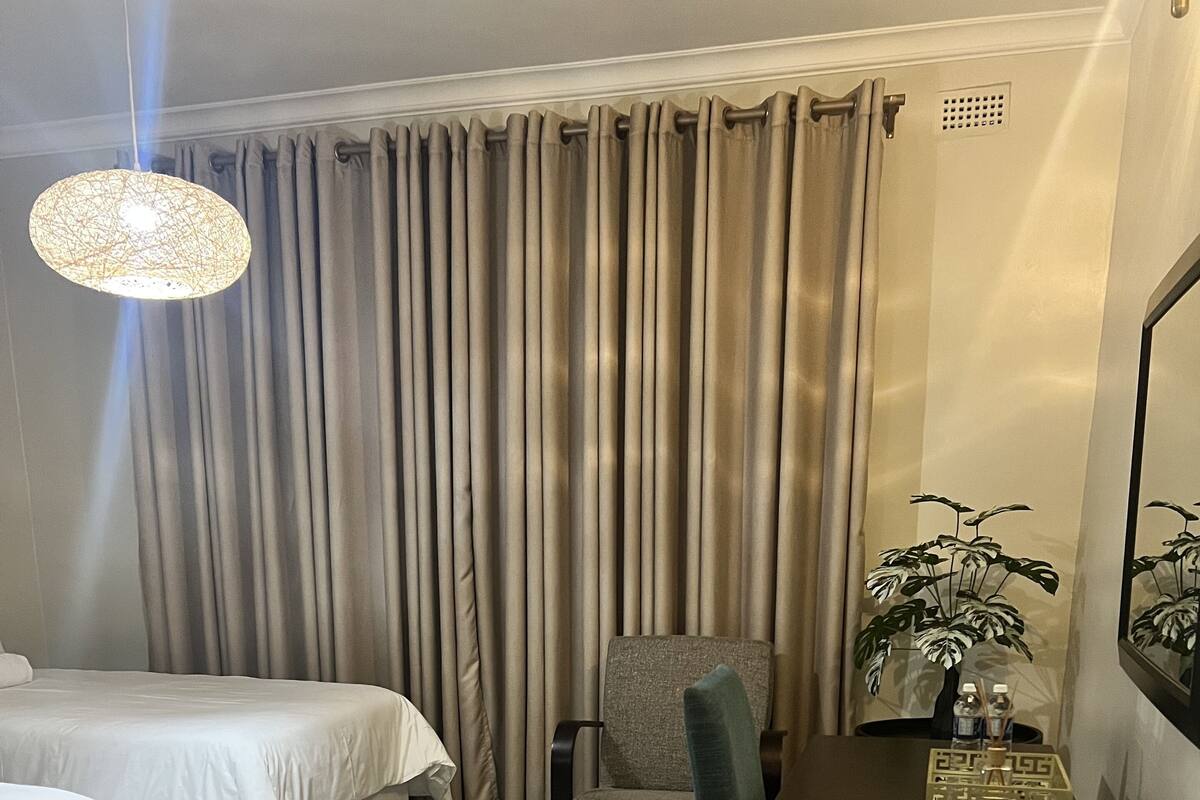 A hotel room with a bed, a chair, a table, a fan, a plant, and a hanging light fixture.