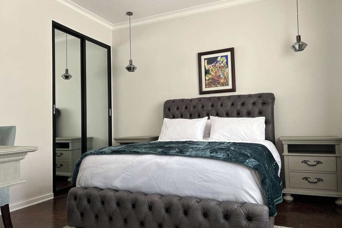 A bedroom with a tufted headboard, a large bed with white and dark blue bedding, a bedside table with drawers, a nightstand, and a framed artwork on the wall.
