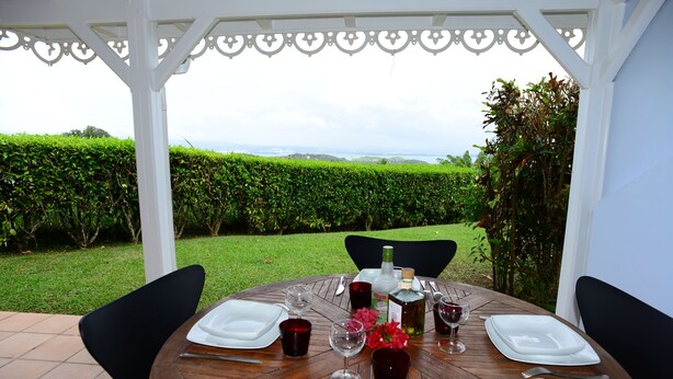 Une table ronde en bois, dressée pour deux personnes, avec de l’argenterie blanche, des couverts et des verres à vin rouge, entourée de chaises noires, sous une pergola blanche offrant une vue sur une haie verdoyante et un paysage lointain.