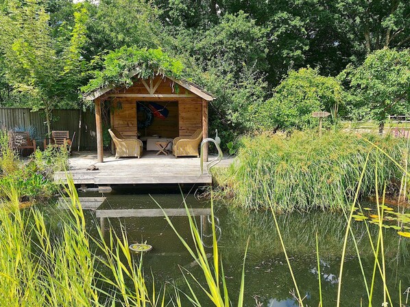 A wooden cabin with a deck, surrounded by lush greenery and a pond.