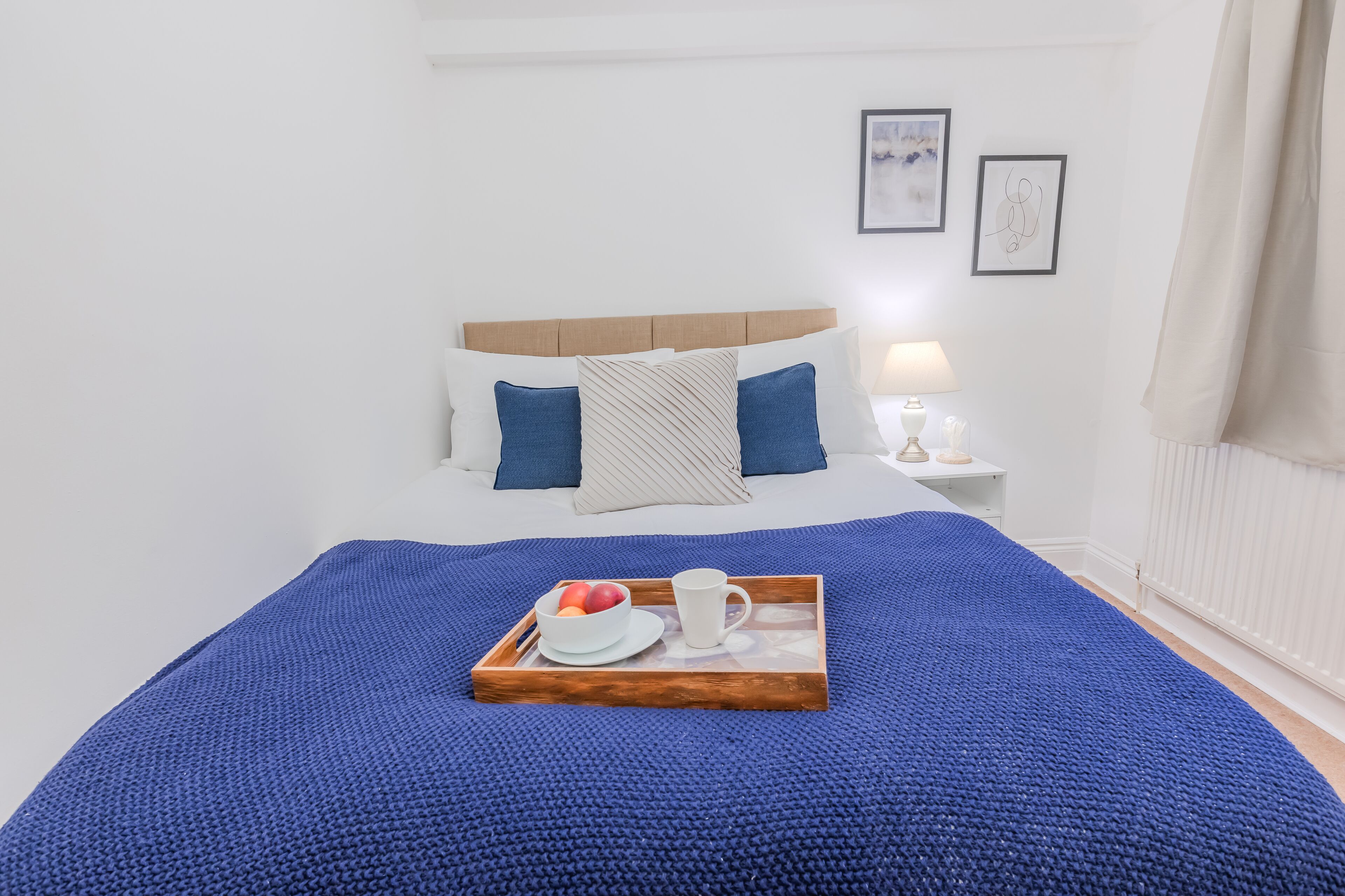 A neatly made bed with a blue blanket, a tray with fruits and a cup, and a bedside table with a lamp.