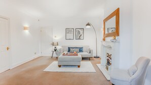 Ein Wohnzimmer mit einem weißen Sofa, einem grauen Hocker, einem weißen Sessel, einem Kamin und zwei gerahmten Bildern an der Wand.