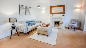 Ein Wohnzimmer mit einem weißen Sofa, einem Couchtisch, einem Beistelltisch, einer Lampe, einem Kamin und einem Sessel.