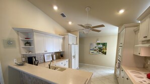 A kitchen with white cabinets, a central island, a ceiling fan, and a painting of a beach scene. - The Island House on Pine Island
with heat-pump heated  most privacy pool (Bokeelia)
