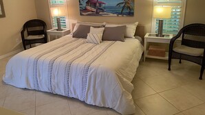 A bedroom with a large bed, two nightstands, two chairs, a painting of a beach scene, and a window with blinds. - The Island House on Pine Island
with heat-pump heated  most privacy pool (Bokeelia)