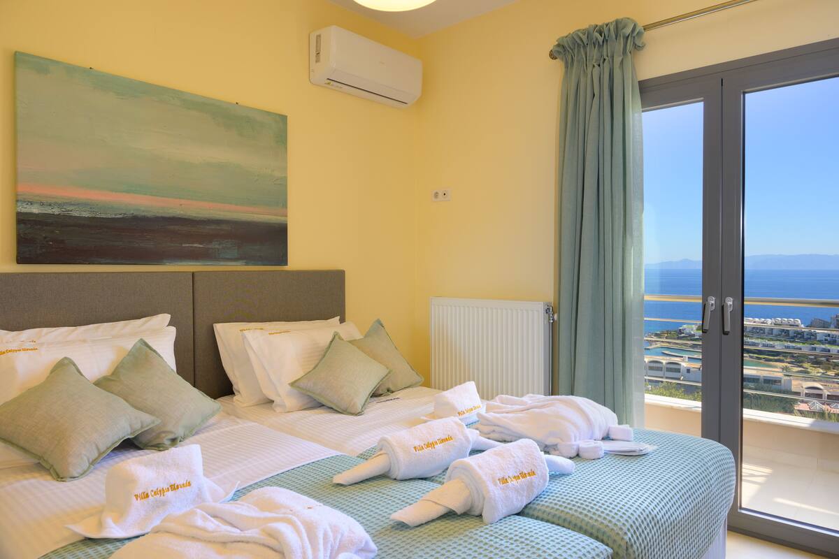 A hotel room with a bed, pillows, towels, an air conditioner, a window with a view, and a painting on the wall.