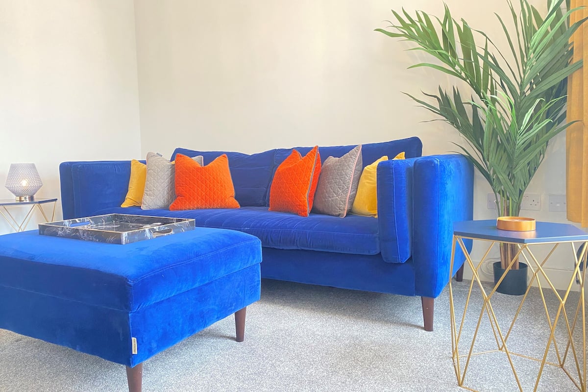 Um sofá azul com várias almofadas, um otomano combinando, uma mesinha com uma planta e um tabuleiro decorativo.