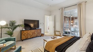A modern bedroom with a bed, a TV, a wooden cabinet, a plant, and a balcony with a view of a brick building. - Spacious 4BR/4.5BA, Pool & Heated Spa, Close Proximity to Dining, Shopping & Fun (Plano)