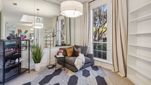 A modern living room with a grey sofa, a side table with a vase, a potted plant, and a large window with curtains. - Spacious 4BR/4.5BA, Pool & Heated Spa, Close Proximity to Dining, Shopping & Fun (Plano)