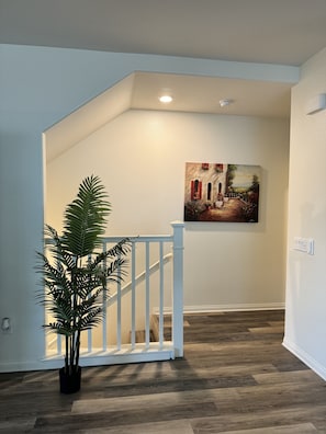 Um corredor com piso de madeira, paredes brancas e corrimão branco. Há uma planta em vaso e um quadro na parede.