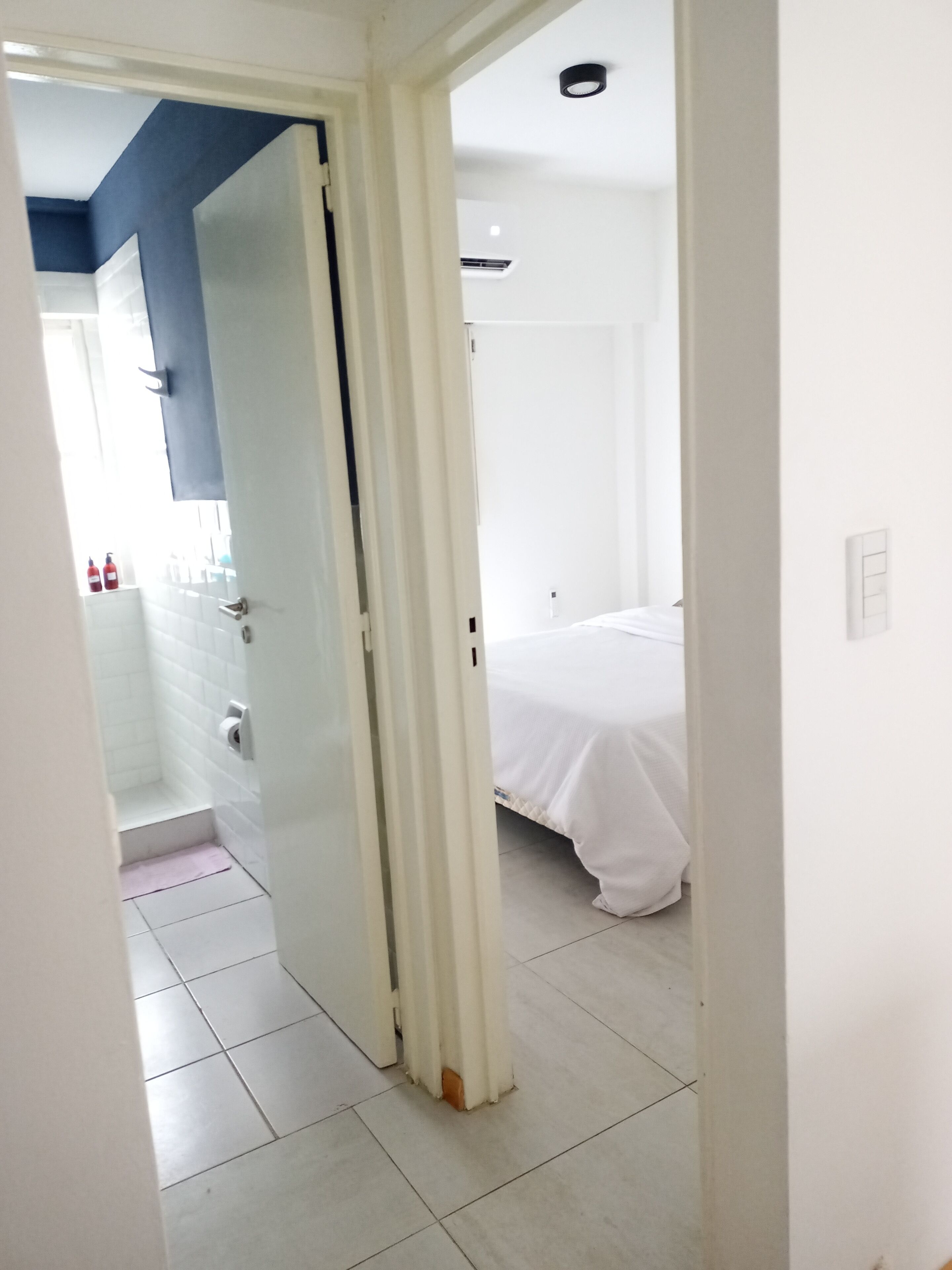 Habitación de hotel con cama blanca, baño con ducha y pasillo que lleva a otra habitación.