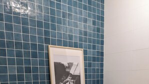 Ein Badezimmer mit blauen Fliesen, einem gerahmten Foto auf einem Holzbrett, einem grünen Strauß, gefalteten Handtüchern und einem gewebten Teppich.