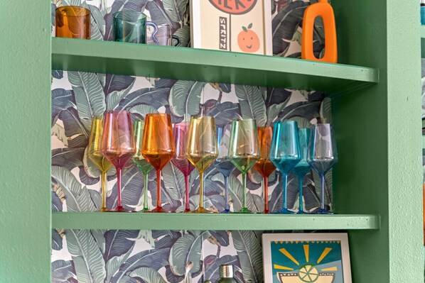 Une étagère avec des verres à vin colorés, le tout placé devant un papier peint orné d’un motif de feuilles.