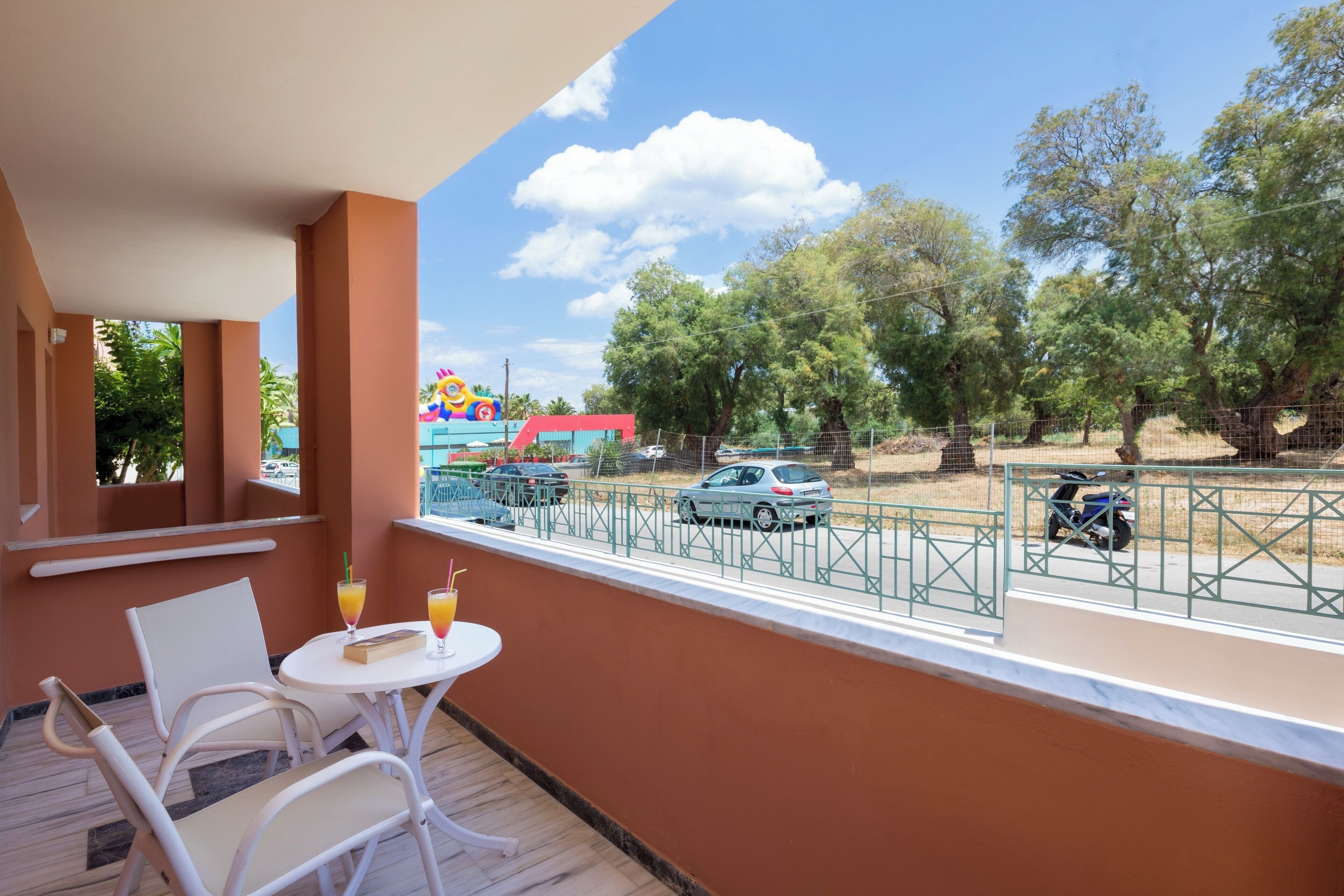 A balcony with white chairs and a small table, overlooking a street with cars and trees.