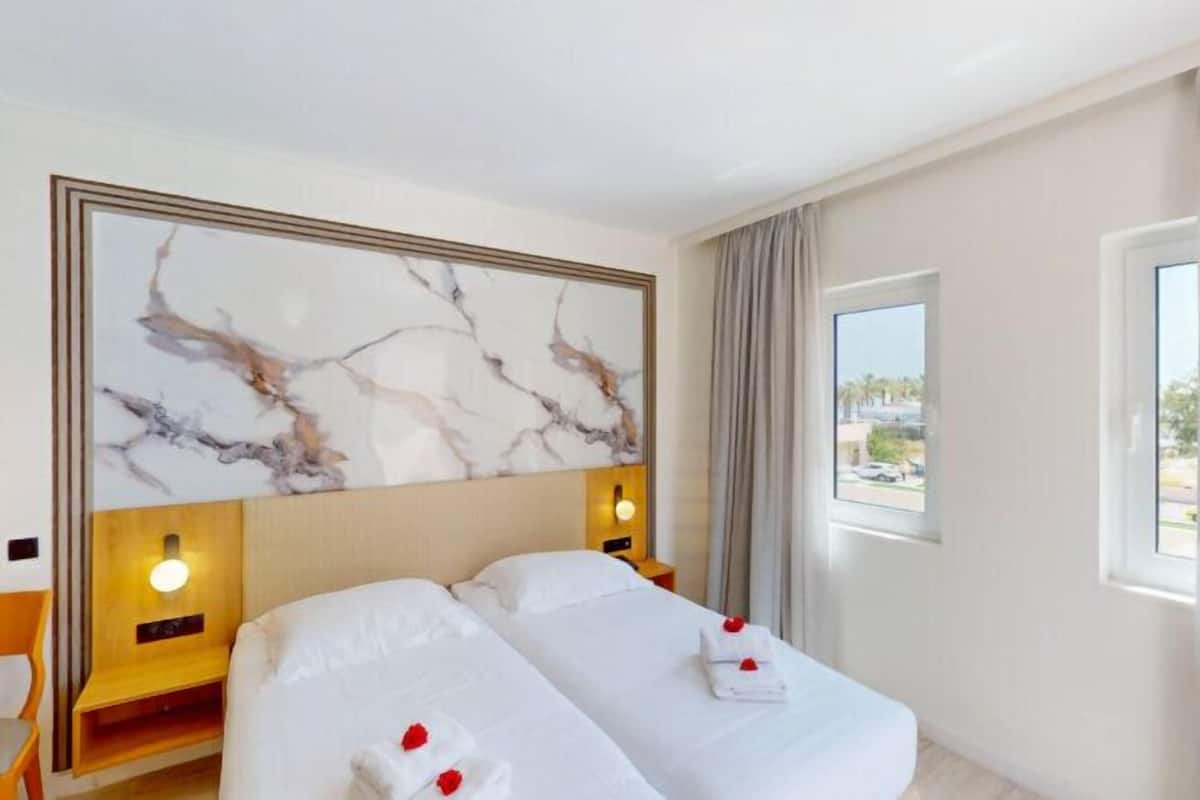 A hotel room with two beds, a marble headboard, bedside lamps, a window with a view, and a wooden chair.