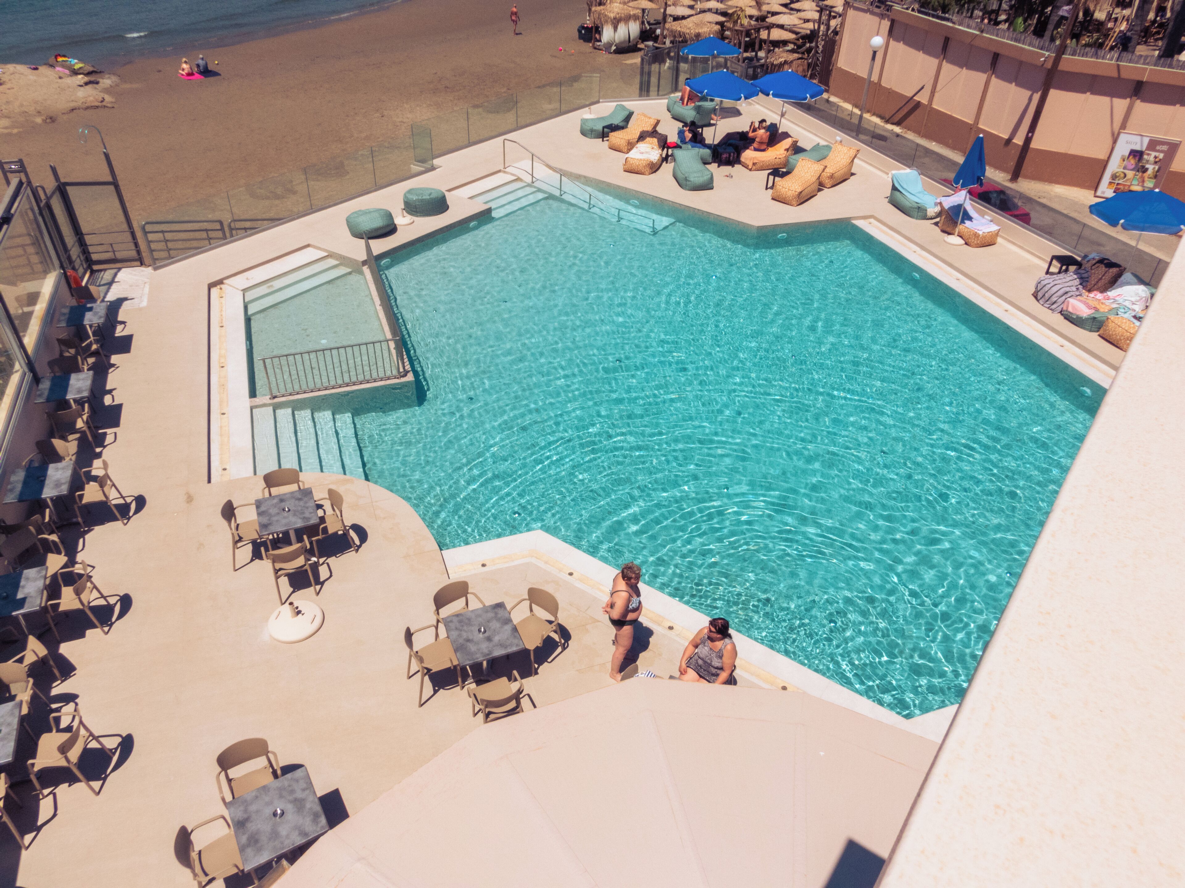 Un área de piscina con sillones reclinables y mesas, una piscina cristalina y una playa al fondo.