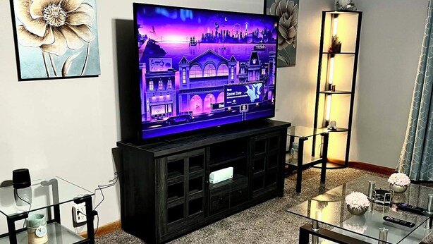 Ein Wohnzimmer mit einem Flachbildfernseher auf einem schwarzen Standfuß, einem Glastisch und einem Teppichboden.