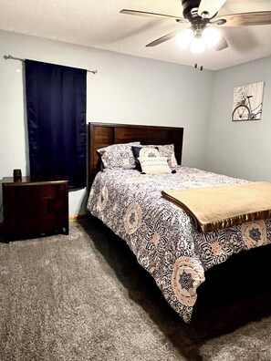 Une chambre à coucher comprenant un lit, une table de chevet, un ventilateur de plafond, une fenêtre avec des rideaux et une œuvre d’art représentant un vélo, accrochée au mur.