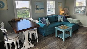 Una sala de estar acogedora con un sofá verde azulado, sillas blancas, una mesa de madera y una pequeña mesa de centro azul. Hay decoraciones de estrellas de mar en los alféizares de las ventanas y un cuadro con motivos playeros en la pared.
