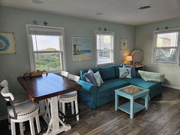 A cozy living room with a teal sofa, white chairs, a wooden table, and a small blue coffee table. There are starfish decorations on the windowsills and a beach-themed painting on the wall.
