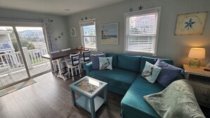 Una sala de estar con un sofá color turquesa, una mesa de centro y un comedor con sillas blancas y una mesa de madera. Hay ventanas con persianas, un balcón y vistas a casas.