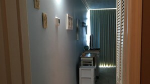 Un pasillo estrecho con paredes azul claro, suelo de baldosas y una puerta a la izquierda. Se observan adornos en la pared y una ventana con persianas en el fondo.